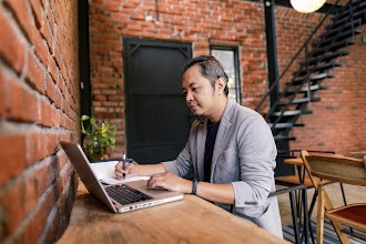 A man photographed from the side is sitting at a table, scrolling on his laptop with one hand, and taking notes in a notebook with the other.