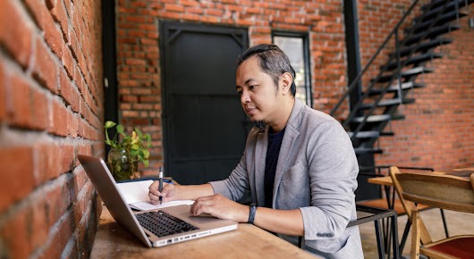 A man photographed from the side is sitting at a table, scrolling on his laptop with one hand, and taking notes in a notebook with the other.
