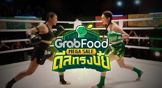 The video thumbnail shows a comedic scene of two people in a Muay Thai boxing ring, one dressed to represent GrabFood, competing in a "Mega Deal Showdown" to promote the GrabFood Mega Sale.
