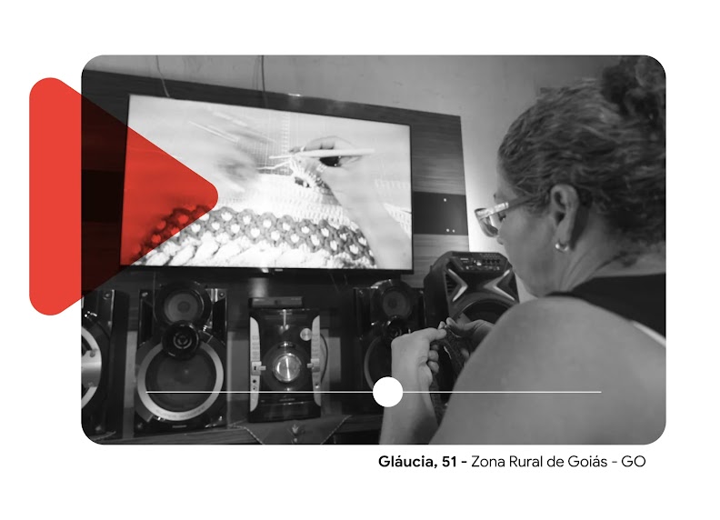Imagem com Gláucia, uma senhora que está fazendo crochê, sentada de frente para a televisão em sua casa. A imagem é em p&b com um símbolo de ‘play’ em vermelho sobreposto.