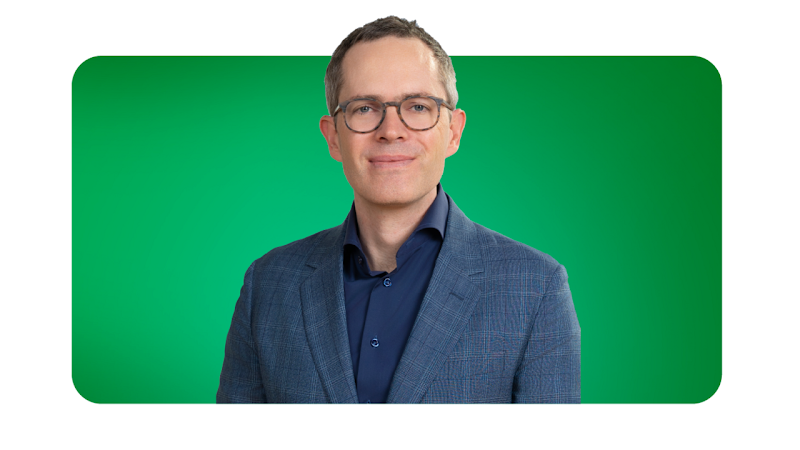 Derek Rodenhausen, managing director e partner do Boston Consulting Group, aparece em frente a um fundo verde. Ele tem pele clara, cabelo curto e castanho, olhos claros; usa óculos e um blazer com padrão xadrez discreto sobre uma camisa escura.