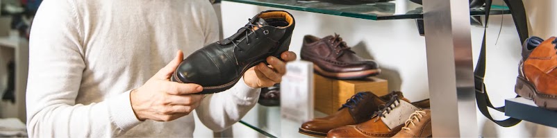 A close-up of a man in a white sweater holding a black leather boot with his hands, inspecting it in a shoe store. Other leather dress shoes in various shades of brown are displayed on glass shelves behind him.