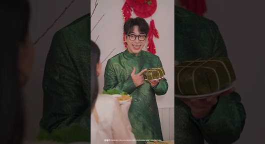 A smiling man in a green traditional Áo Dài points to a square Bánh Chưng cake he is holding on a plate, with red Lunar New Year decorations in the background