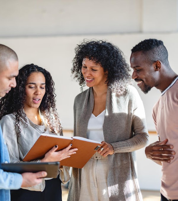 Two women and two men are standing next to each other, looking at the notebook that the woman on the left is holding. The man on the left is holding a tablet.