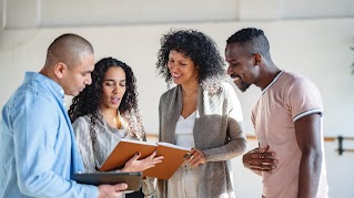 Two women and two men are standing next to each other, looking at the notebook that the woman on the left is holding. The man on the left is holding a tablet.