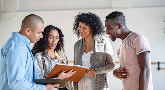 Two women and two men are standing next to each other, looking at the notebook that the woman on the left is holding. The man on the left is holding a tablet.