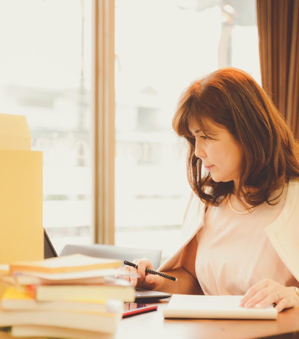 A woman sitting at her desk is looking at her laptop, which is partly hidden by a pile of books and folders. She has a pencil in one hand, and her other hand lies on a notebook.