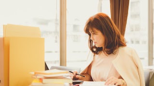 A woman sitting at her desk is looking at her laptop, which is partly hidden by a pile of books and folders. She has a pencil in one hand, and her other hand lies on a notebook.