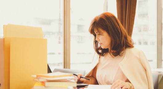 A woman sitting at her desk is looking at her laptop, which is partly hidden by a pile of books and folders. She has a pencil in one hand, and her other hand lies on a notebook.