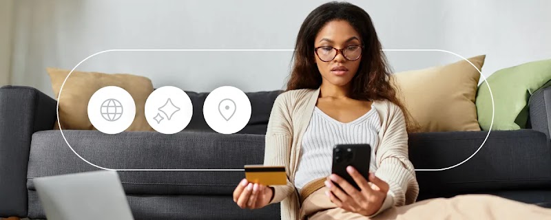 A woman with long hair and glasses is sitting on a sofa. She holds a mobile phone in one hand, and a credit card in the other. She is looking at the phone screen. To the left, 3 icons: an internet icon; the Gemini icon; and a location pin.