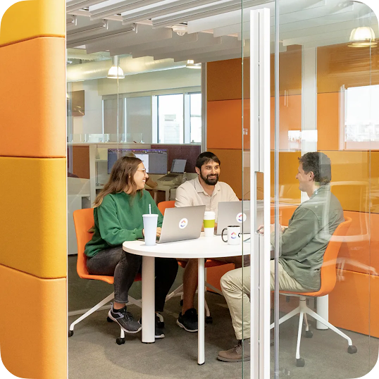 Three ClickUp colleagues sitting at a white round table in a modern, brightly colored office space, engaged in a collaborative meeting. A stat overlay reads “+16% return on ad spend.”
