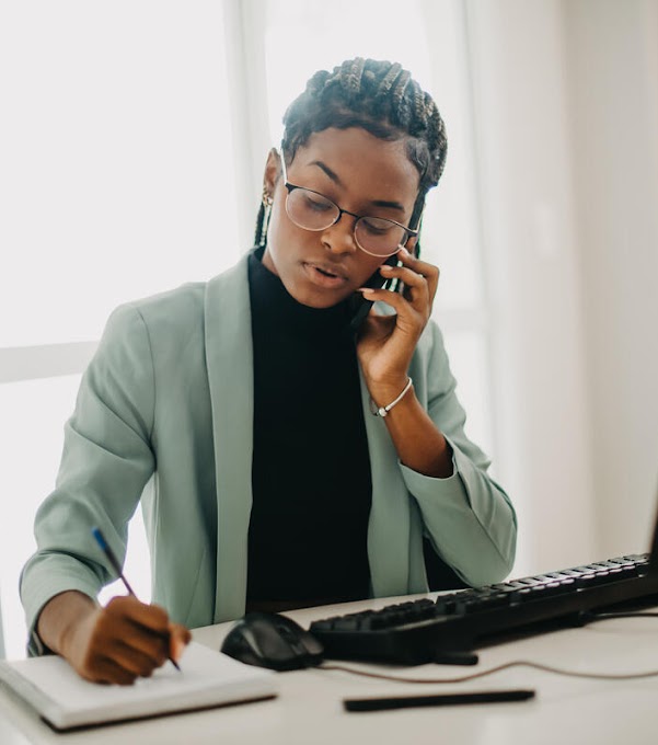A woman with a green suit and glasses is sitting at a desk in front of a monitor. She is on the phone and writing in a notebook.