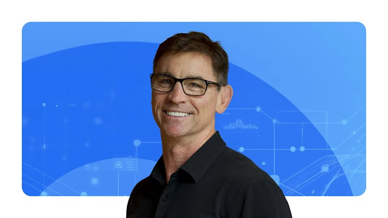 Paul Limbrey, Google’s VP of global 3P ecosystem, appears in front of a background of blue arcs. Limbrey has light skin, dark hair, and wears glasses and a black collared shirt.