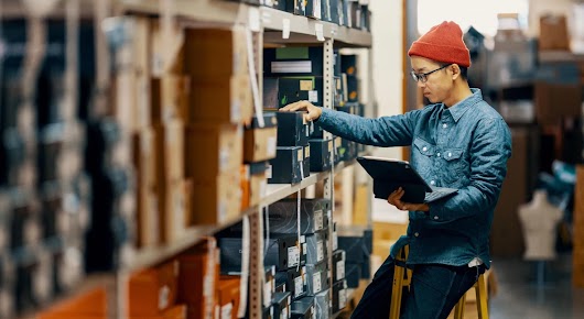A retail worker manages their store inventory, looking through shelves with laptop in hand.