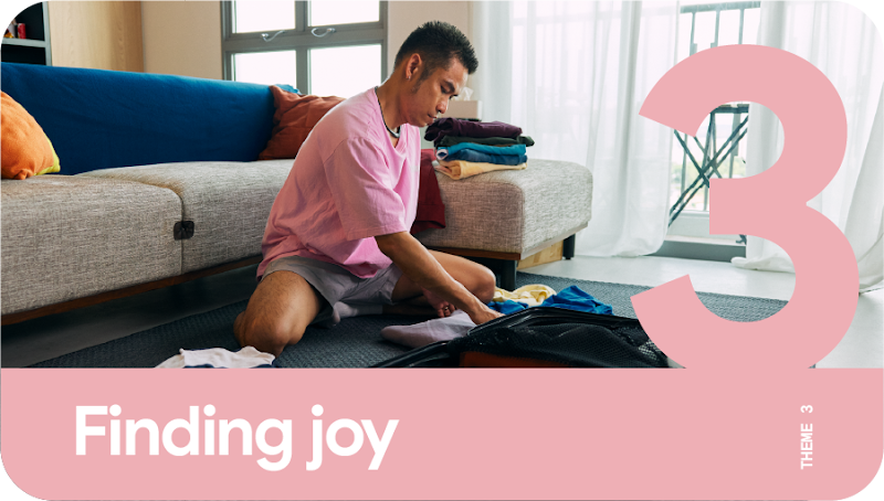 Theme 3: Finding joy. A man with a pink shirt sitting cross-legged in front of a gray sofa, folding his laundry on a carpeted floor.