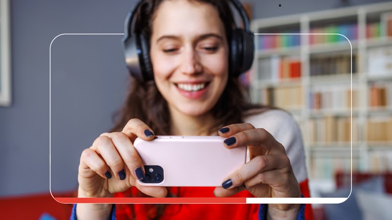 Donna con pelle chiara e capelli lunghi scuri con maglia rossa e bianca, smalto nero alle unghie e cuffie over-ear di colore nero. Tiene in mano un cellulare con cover rosa con cui guarda video di YouTube sui suoi giochi preferiti.