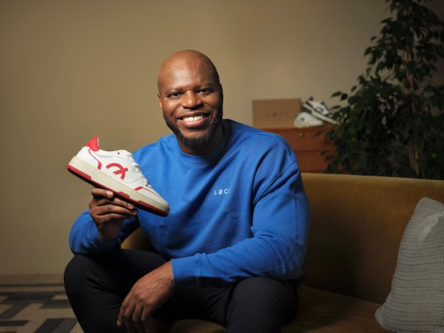   A man with a shaved head and a beard sits on a tan colored couch, smiling and holding up a white sneaker with red accents. He wears a bright blue long-sleeved shirt with a small white logo on the chest. In the background, there is a wooden dresser with a shoebox and another pair of sneakers, along with a potted plant.