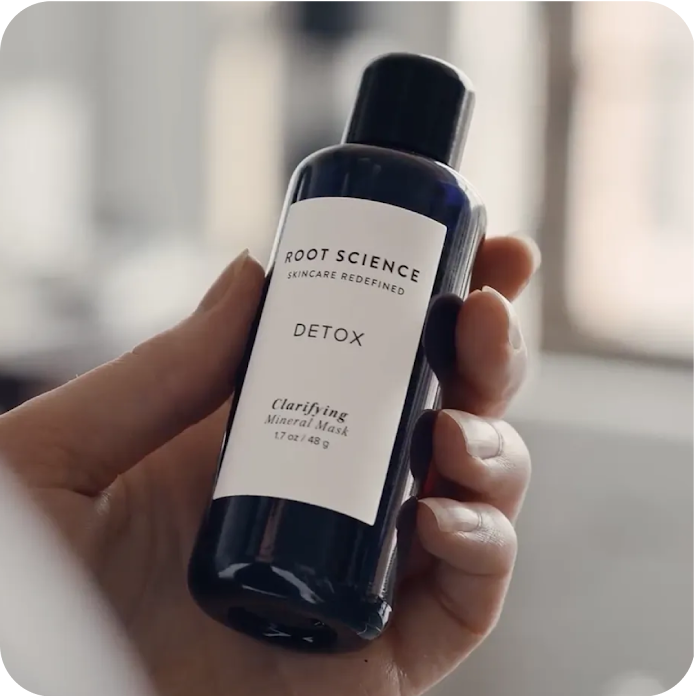 Sebuah tangan memegang botol biru kecil yang merupakan produk masker mineral Root Science.
