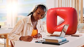 A young woman with headphones takes notes at a desk beside a laptop and a large red inflatable YouTube play button.
