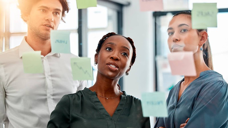 Un equipo de tres jóvenes profesionales, un hombre de piel clara, una mujer de piel negra y otra mujer de piel media, analizan concentrados varias notas adhesivas de colores pegadas en un cristal en una oficina.