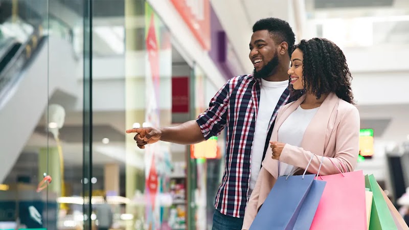 A Black couple is shopping at a mall. The man, wearing a plaid shirt, points at a store window, smiling at the woman next to him. She holds several colorful shopping bags and looks in the direction he's pointing, smiling.