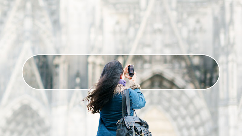 A woman with long black hair and a blue jacket stands with her back to the viewer. She is taking a picture of an ornate cathedral in front of her with her phone. There is an overlay of a Google Search bar over the cathedral.