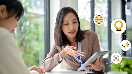 Two women in discussion while looking at an iPad with a globe, location, loudhailer and magnifying glass with a dollar sign surrounding them, representing how Spark X uses AI marketing to help exporters expand globally and increase ROI and profits.