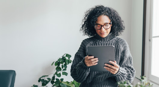 グレーのセーターを着た女性が手に持ったタブレットを見ている。女性の後ろには観葉植物、右手側には窓がある。