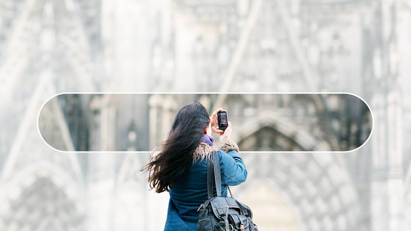 Une femme aux longs cheveux noirs et portant une veste bleue se tient de dos. Elle prend en photo avec son téléphone une cathédrale richement décorée. La barre de recherche Google est superposée à l'image de la cathédrale.