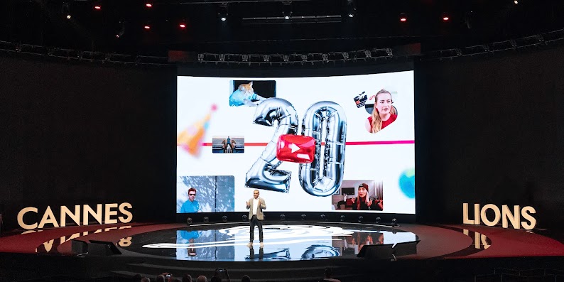 Neal Mohan, CEO di YouTube, parla da un palco ovale lucido al Cannes Lions Festival davanti a un grande schermo con una slide che mostra screenshot di video di YouTube e palloncini in Mylar a forma di 20 e dell'icona del pulsante di riproduzione.