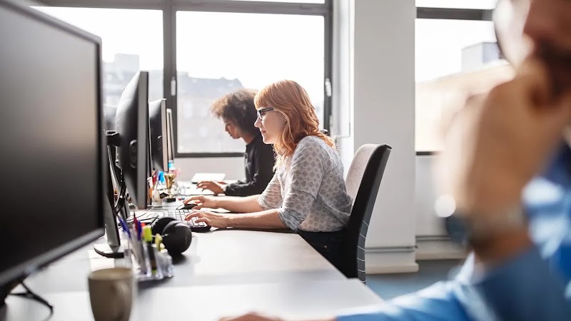 Una mujer con cabello rubio y gafas se sienta entre dos compañeros de trabajo y usa una computadora de escritorio. La fila de escritorios está junto a una gran ventana.