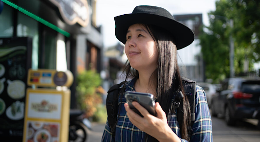 A woman wearing a hat is walking past a restaurant, phone in hand.