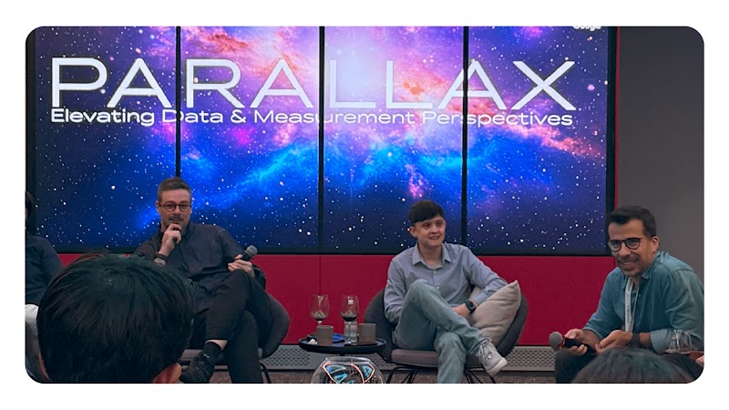 Três homens sentados em poltronas durante um painel de discussão, com microfones na mão e copos sobre a mesa. Ao fundo, uma tela grande exibe o título ‘PARALLAX – Elevating Data & Measurement Perspectives’, com uma imagem de galáxia colorida.