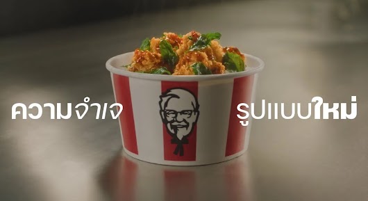 A KFC bucket filled with spicy, herb-topped fried chicken, with Thai text on the left reading "ความจำเจ" and on the right reading "รูปแบบใหม่"