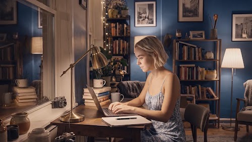 A young blonde woman sitting at a wooden desk, typing on a laptop in a cozy, softly lit room at night.