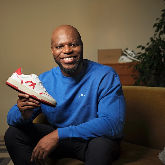 رجل ذو رأس حليق ولحية يجلس على أريكة بلون بيج (تان)، وهو يبتسم ويمسك بحذاء رياضي أبيض مع لمسات باللون الأحمر. يرتدي قميصاً أزرق زاهياً بأكمام طويلة مع شعار أبيض صغير على الصدر. في الخلفية، تظهر خزانة خشبية عليها صندوق أحذية وزوج آخر من الأحذية الرياضية، بالإضافة إلى نبتة منزلية.