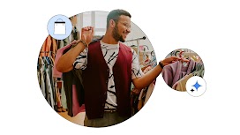 Un hombre de tono de piel media y pelo negro, con gafas y sonriente, lleva un chaleco granate y elige ropa en una tienda. Sostiene una prenda en una percha mientras mira otras opciones en un perchero.