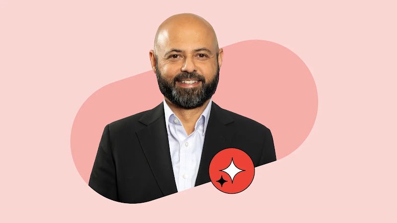 Headshot of Mustafa Peracha, Chief Consumer Officer at Ooredoo Qatar, smiling with a beard and shaved head, wearing a dark suit and open-collar shirt against a pink background with a red sparkle icon.