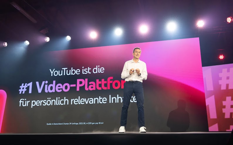 Philipp Justus, VP Central Europe bei Google, steht auf einer Bühne. Im Hintergrund ist auf einem großen Bildschirm der Text „YouTube ist die #1 Video-Plattform für persönlich relevante Inhalte“ zu sehen.