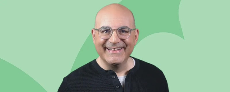 Shelly Palmer, CEO of The Palmer Group and advanced media professor in residence at Syracuse Univ. smiles at the camera, wearing a black pullover and glasses. He has light skin and a shaved head.