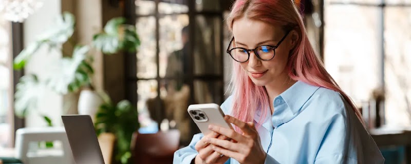 A woman with long blonde hair and pink streaks is sitting at a desk. She is wearing glasses and a pale blue shirt. An open laptop and a coffee cup sit in front of her. She is holding a mobile phone in both hands, and is looking at the screen.