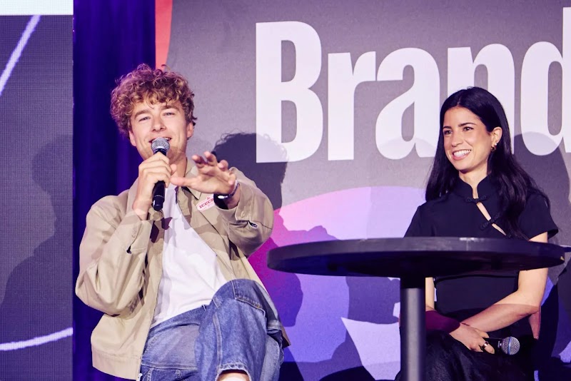 Le journaliste et créateur de contenu Gaspard G, micro en main, intervient sur la scène de l'événement « Creator Collective ». À ses côtés, la créatrice Stelle l'écoute devant un arrière-plan affichant le mot « Brand ».