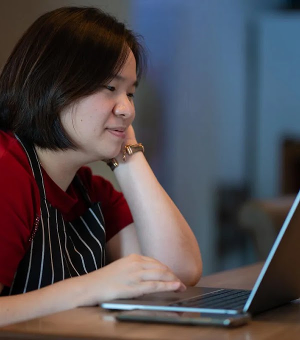 Mujer propietaria de un pequeño negocio, con delantal, sentada en un escritorio trabajando en una computadora portátil.