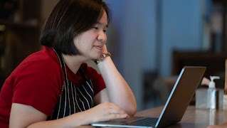Mujer propietaria de un pequeño negocio, con delantal, sentada en un escritorio trabajando en una computadora portátil.