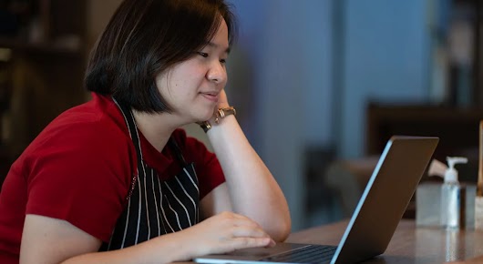 Mujer propietaria de un pequeño negocio, con delantal, sentada en un escritorio trabajando en una computadora portátil.