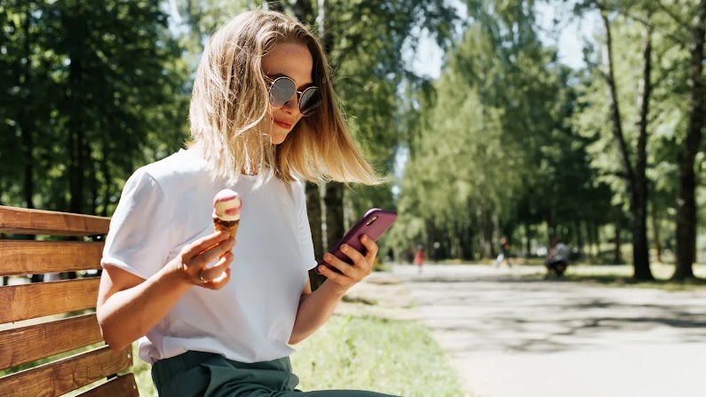 A woman with shoulder-length blonde hair and wearing a white T-shirt is sitting on a park bench. She is wearing sunglasses, and is holding an ice-cream cone in her right hand, and a mobile phone in her left hand. She is looking at the phone screen.