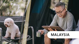 Malaysia. An older man sitting in a tent holding a cup in one hand and a tablet, with a dog on the seat beside him.