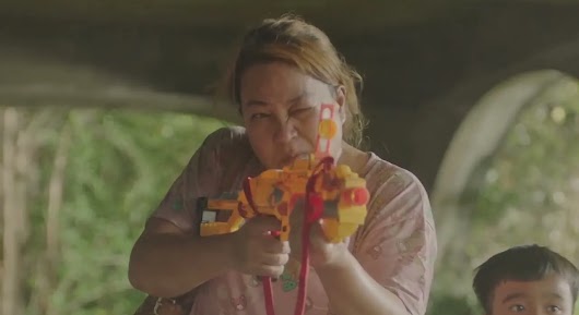 A woman with one eye closed aiming a bright yellow and orange toy gun directly at the camera.