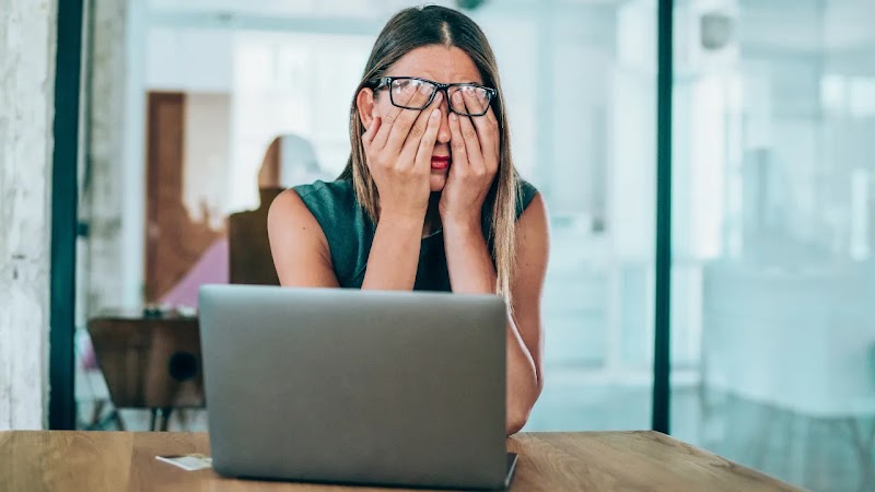 A stressed woman with glasses sitting at a desk in front of a laptop, covering her eyes and face with her hands in frustration.