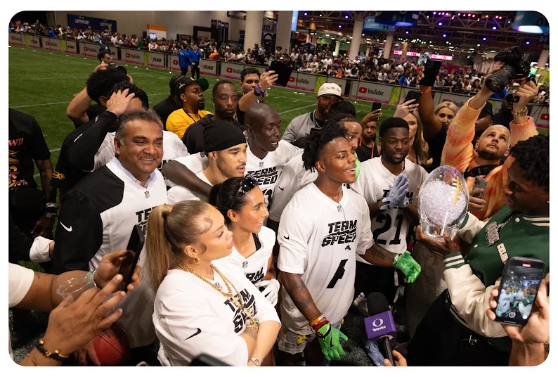 A group of YouTube creators wearing shirts that say “Team Speed” gather on an indoor football field around a crystal, football-shaped trophy.